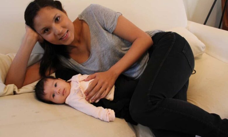 A woman in a grey t-shirt and black pants is lying on a bed and looking into the camera as she's put her hand on an infant's belly