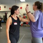 Two women push their left hands together as a form of exercise during the Power Hour CrossFit class. Both women have ponytails in their hair and they're wearing gym workout clothing.