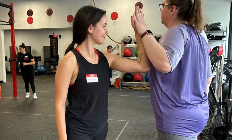 Two women push their left hands together as a form of exercise during the Power Hour CrossFit class. Both women have ponytails in their hair and they're wearing gym workout clothing.