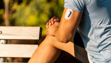 A young man outdoors stretching his leg on a bench wears a continuous glucose monitor on his upper arm.