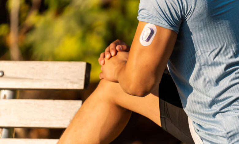 A young man outdoors stretching his leg on a bench wears a continuous glucose monitor on his upper arm.